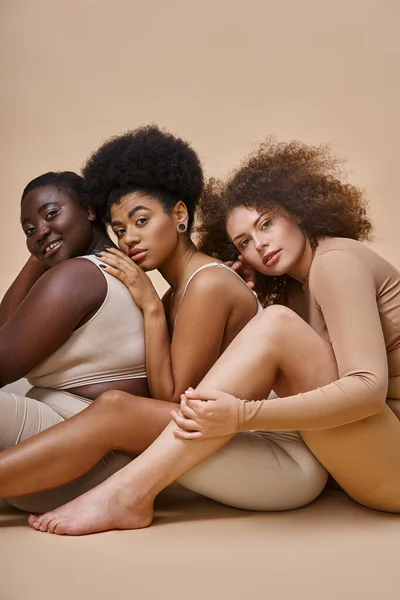 Ausdrucksstarke körperpositive Frauen in Unterwäsche sitzend und in die Kamera schauend auf beige, Selbstbewusstsein — Stockfoto