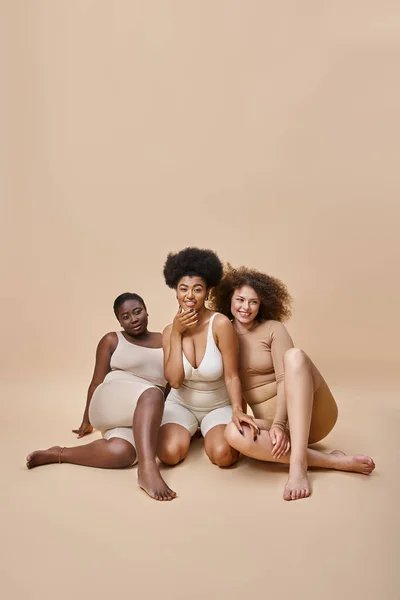 Tres alegres mujeres multirraciales más tamaño en ropa interior sentado en beige, belleza natural del cuerpo positivo - foto de stock