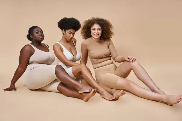 Multicultural más tamaño de las mujeres en ropa interior sentado y mirando a la cámara en gris, además de belleza tamaño - foto de stock