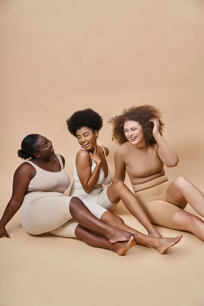 Sorridente multiétnico plus size modelos em roupa interior sentado em pano de fundo cinza, plus size positivity — Fotografia de Stock