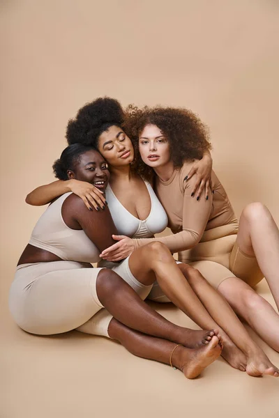 Amigos femeninos multiétnicos de talla grande en ropa interior sentados y abrazados en gris, juntos - foto de stock