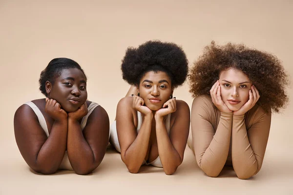 Encantador cuerpo multiétnico mujeres positivas en lencería acostado y mirando a la cámara en beige - foto de stock