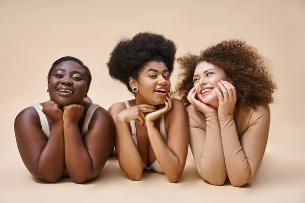 Sonriente cuerpo multiétnico mujeres positivas en lencería acostado y posando en beige, belleza natural - foto de stock