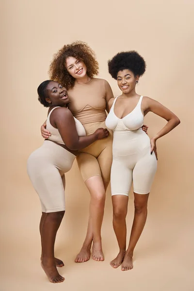 Comprimento total de mulheres multirraciais alegres em roupa interior posando em bege, beleza e positividade corporal — Fotografia de Stock