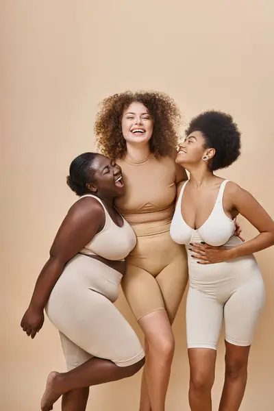 Mulheres multirraciais alegres em roupa interior posando em pano de fundo bege, beleza e positividade corporal — Fotografia de Stock