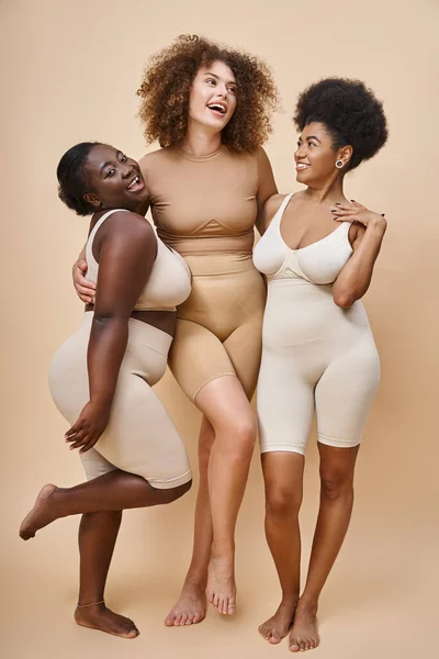 Longitud completa de alegres mujeres multirraciales en ropa interior posando en beige, belleza y positividad corporal - foto de stock
