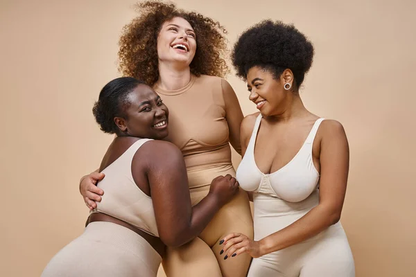 Fröhliche multiethnische Frauen in Unterwäsche posieren auf beigem Hintergrund, Schönheit und Körperpositivität — Stock Photo