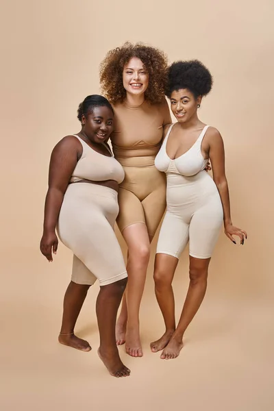 Comprimento total de mulheres multirraciais alegres em roupa interior posando em bege, beleza e positividade corporal — Fotografia de Stock