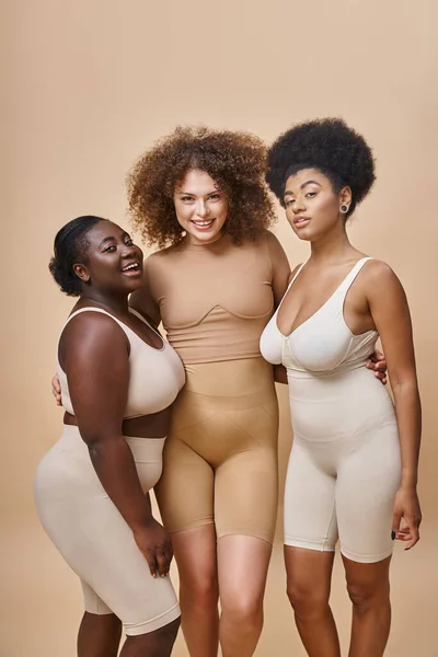 Heureux multiethnique plus la taille copines en lingerie posant sur fond beige, corps beauté positive — Photo de stock