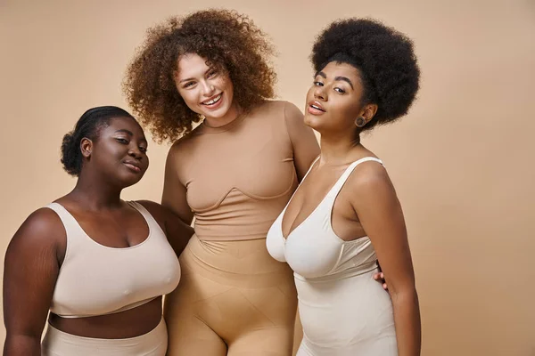 Glücklich multiethnische plus size Freundinnen in Dessous posieren auf beigem Hintergrund, Körper positive Schönheit — Stockfoto