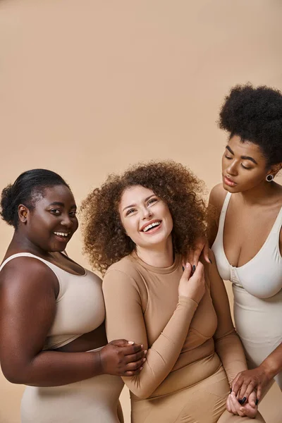 Joyeuses petites amies multiethniques plus de taille en lingerie posant sur fond beige, positivité du corps — Photo de stock