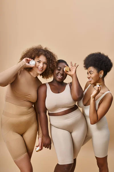 Glückliche multiethnische Plus-Size-Frauen in Dessous posieren mit Gläschen kosmetischer Creme auf beige, Hautpflege — Stockfoto