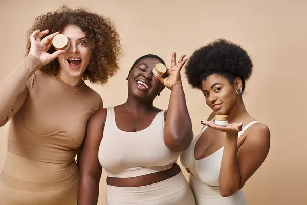 Happy multiethnic plus size women in lingerie posing with jars of cosmetic cream on beige, skincare — Stock Photo