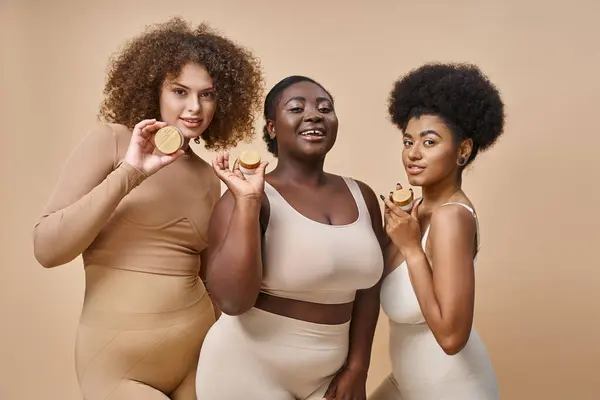 Happy multiethnic plus size women in lingerie posing with jars of cosmetic cream on beige, skincare — Stock Photo