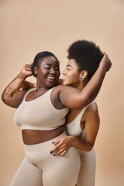 Cheerful african american girlfriends in lingerie having fun while posing on beige, curvy beauty — Stock Photo