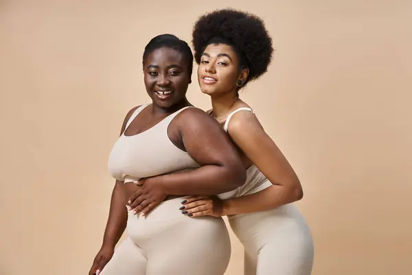 Encantador plus size mulheres americanas africanas em roupa interior olhando para a câmera em bege, positividade do corpo — Fotografia de Stock