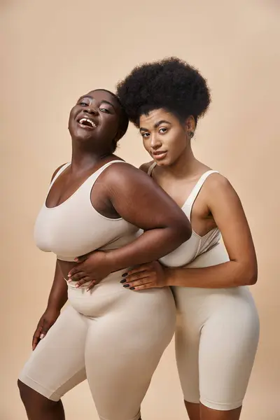 Радостный плюс размер африканских американских женщин в нижнем белье, глядя на камеру на бежевый, тело позитивности — стоковое фото