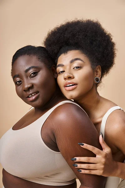 Gioiosa plus size donne africane americane in biancheria intima guardando la fotocamera sul beige, positività del corpo — Foto stock