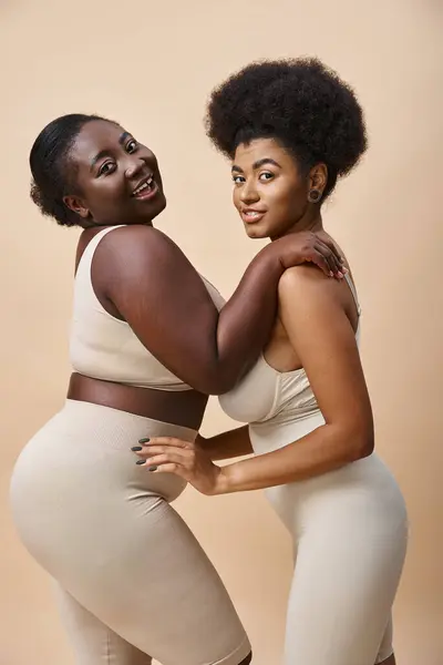 Sensual and smiling plus size african american women posing in lingerie on beige, curvy beauty — Stock Photo