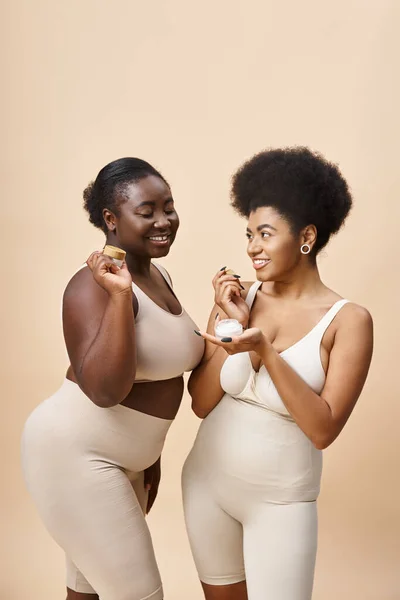 Happy african american plus size women in lingerie holding cosmetic cream on beige, skincare — Stock Photo