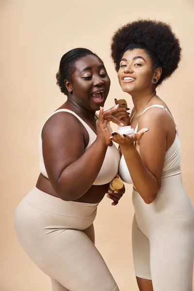Joyeux afro-américain plus les femmes de taille en lingerie posant avec crème cosmétique sur beige, soins de la peau — Photo de stock