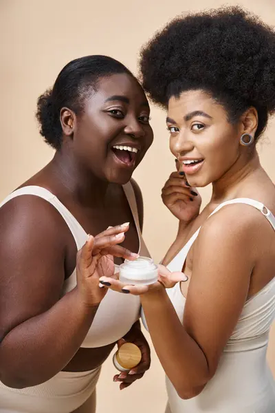 Alegre africano americano más tamaño mujeres en lencería posando con crema cosmética en beige, cuidado de la piel - foto de stock