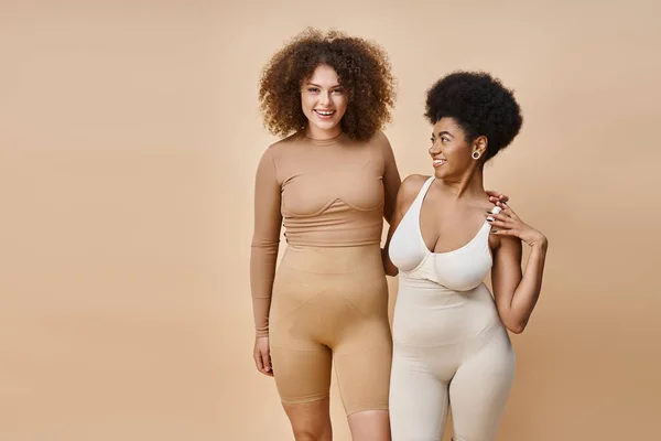 Sorrindo multicultural corpo positivo mulheres em roupa interior de pé em bege, natural plus size beleza — Fotografia de Stock