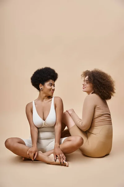 Cheerful multicultural plus size girlfriends in lingerie sitting on beige backdrop, natural beauty — Stock Photo