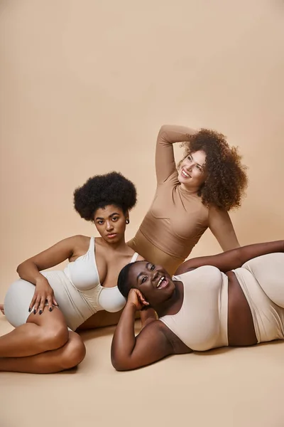 Feliz cuerpo multirracial mujeres positivas en lencería posando sobre fondo beige, belleza natural - foto de stock