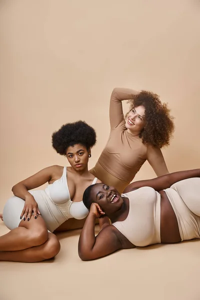 Happy multiracial body positive women in lingerie posing on beige backdrop, natural beauty — Stock Photo