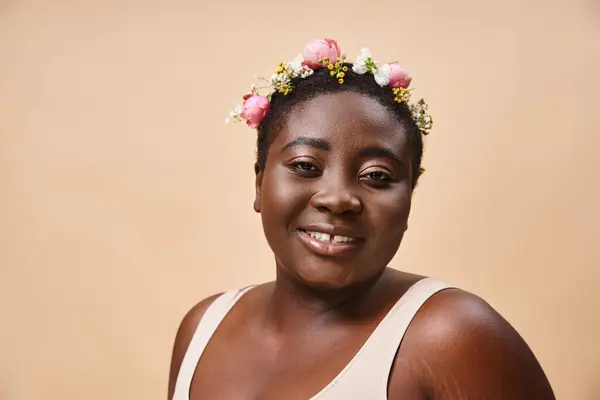Portrait of curvy and happy african american woman with flowers in hair looking at camera on beige — Stock Photo