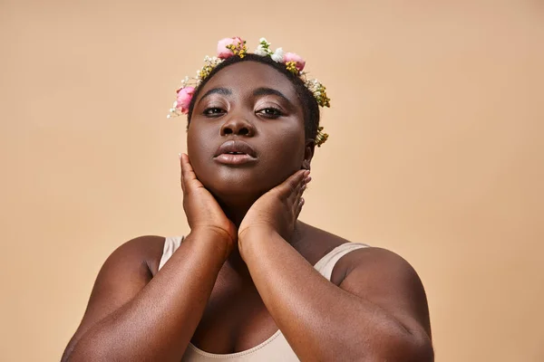 Charmante femme afro-américaine de taille plus avec des fleurs dans les cheveux et les mains près du visage sur beige — Photo de stock