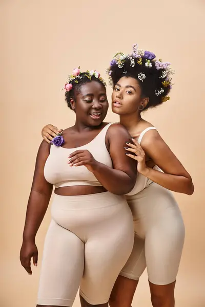Lächelnde afrikanisch-amerikanische Frauen mit Blumen im Haar posieren in Unterwäsche auf beige, plus size beauty — Stockfoto