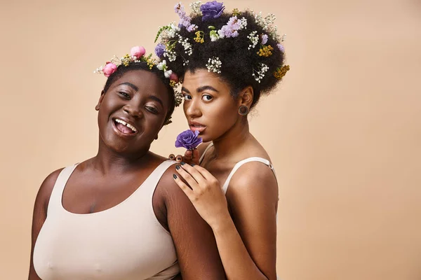 Smiling african american women with flowers in hair posing in underwear on beige, plus size beauty — Stock Photo