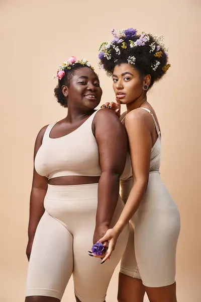 Mujeres afroamericanas encantadoras con flores en el pelo posando en ropa interior en beige, además de belleza de tamaño - foto de stock
