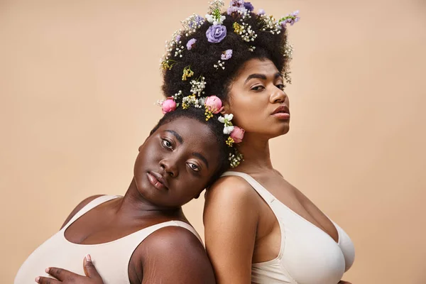 Charmante afrikanisch-amerikanische Frauen mit Blumen im Haar posieren in Unterwäsche auf beige, plus size beauty — Stockfoto
