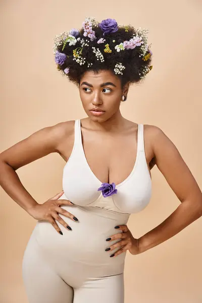Mujer afroamericana confiada con flores en el pelo posando en lencería con las manos en las caderas en beige - foto de stock
