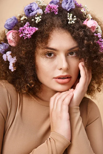 Portrait of charming woman with flowers in wavy hair looking at camera on beige, natural beauty — Stock Photo