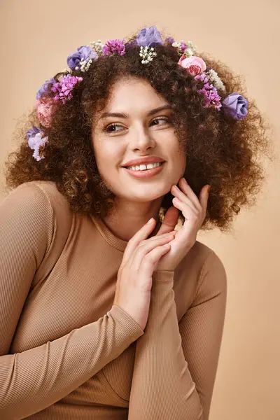 Joyeux jeune femme avec des fleurs colorées dans les cheveux ondulés regardant loin sur beige, beauté naturelle — Photo de stock