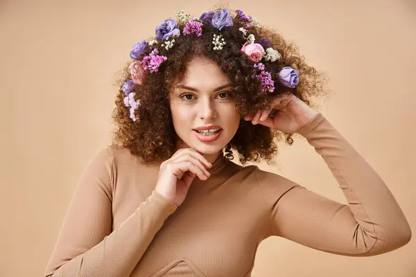 Heureuse jeune femme avec des fleurs colorées en cheveux ondulés regardant la caméra sur beige, beauté naturelle — Photo de stock