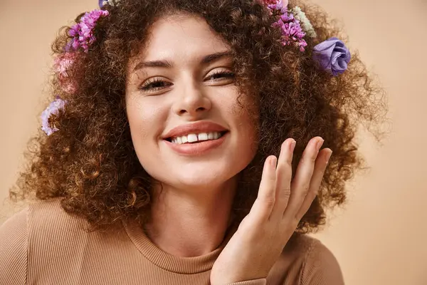 Retrato de mujer feliz con flores de colores en el pelo sonriendo a la cámara en beige, belleza natural - foto de stock