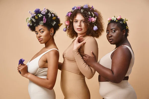 Corps multiethnique femmes positives en lingerie avec des fleurs colorées dans les cheveux sur beige, plus la beauté de taille — Photo de stock