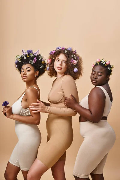 Multiethnic body positive women in lingerie with colorful flowers in hair on beige, plus size beauty — Stock Photo