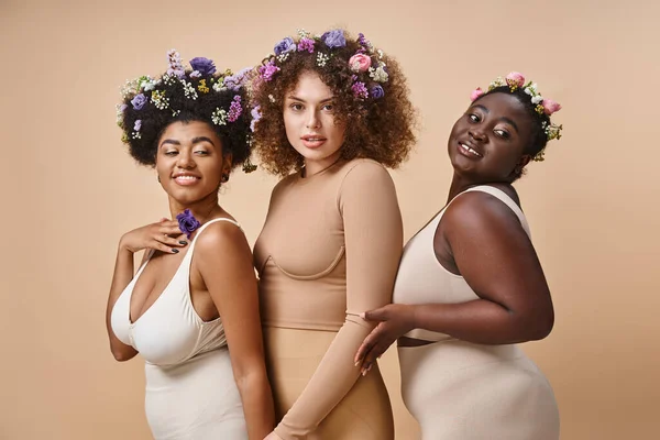 Multiethnische Plus-Size-Frauen in Dessous mit bunten Blumen im Haar auf beige, kurvige Eleganz — Stockfoto