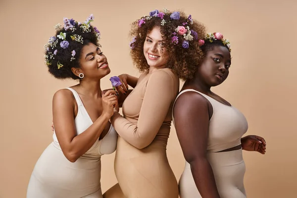 Happy multiracial plus size women in lingerie with colorful flowers in hair on beige, curvy charm — Stock Photo