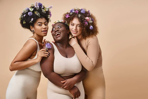 Happy multiracial plus size women in lingerie with colorful flowers in hair on beige, curvy charm — Stock Photo