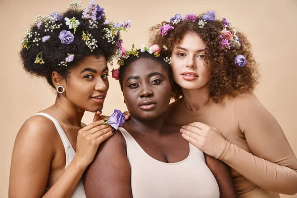 Seductoras mujeres multirraciales en lencería con flores de colores en el pelo en beige, además de belleza de tamaño - foto de stock