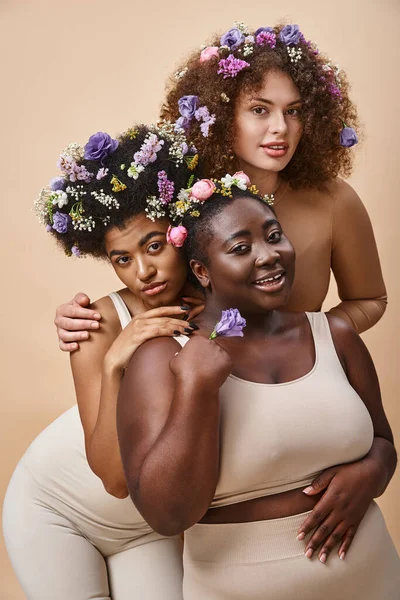 Seductoras mujeres multirraciales en lencería con flores de colores en el pelo en beige, además de belleza de tamaño - foto de stock