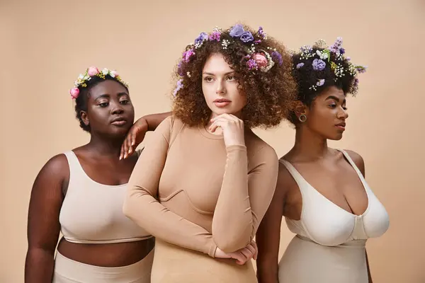 Mujeres multiétnicas de ensueño en lencería con flores de colores en el pelo en beige, además de belleza de tamaño - foto de stock
