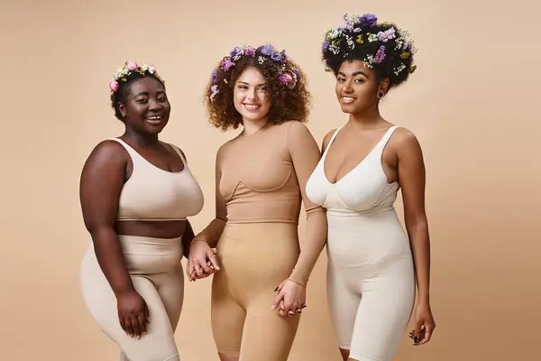 Joyful multiethnic girlfriends in lingerie with colorful flowers in hair on beige, plus size beauty — Stock Photo